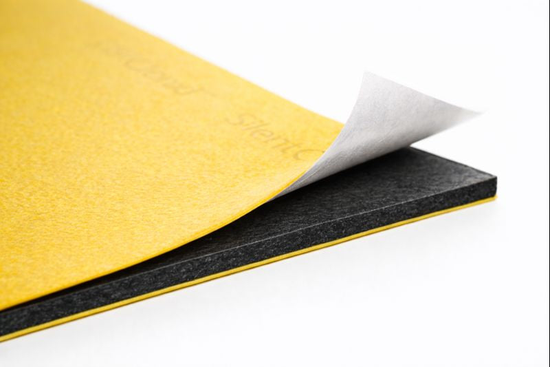 Close-up of SilentCloud ProMass 50 acoustic membrane: yellow top layer with peel-and-stick backing revealing adhesive on black core (2.6 mm, 5 kg/m²) for high-density soundproofing.