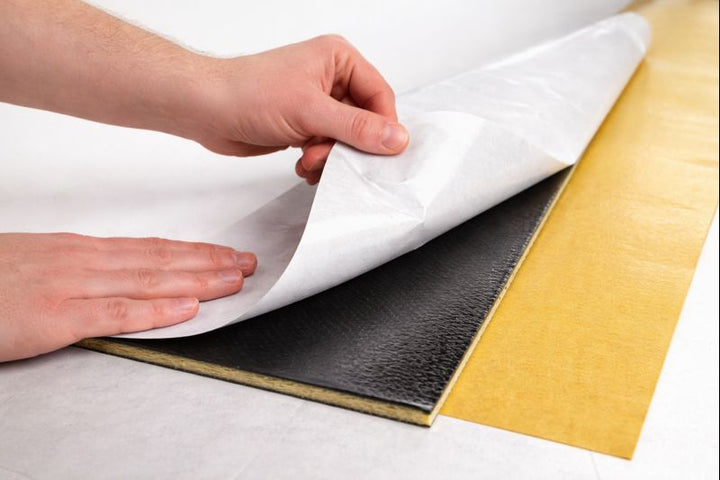Professional showing SilentCloud ProMass 50 peel-and-stick membrane being applied: removing liner to reveal adhesive on black layer with yellow backing (2.6 mm, 5 kg/m²), for walls & ceilings.